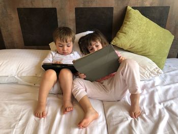 High angle view of siblings using digital tablet while lying on bed at home