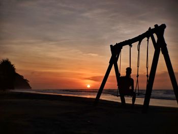 Silhouette man sitting on swing at beach against sky during sunset