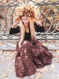 Full length of smiling woman with maple leaves sitting by fence