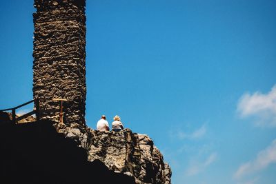 Low angle view of couple sitting by column on cliff against blue sky