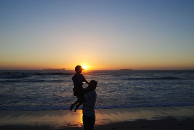 Father carrying son at beach against sky during sunset