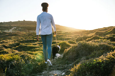 Full body back view of ethnic woman with border collie dog walking together on trail among grassy hills in sunny spring evening