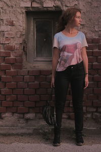 Full length of thoughtful young woman standing against brick wall