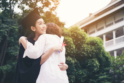 Cheerful woman in graduation gown embracing mother while standing outdoors