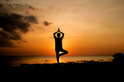 Silhouette person exercising yoga at beach against sky during sunset