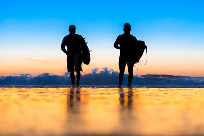 Rear view of men carrying equipment while walking towards sea against sky during sunset