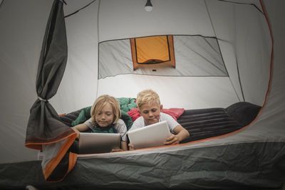 Siblings using digital tablet while lying together in tent at camping site