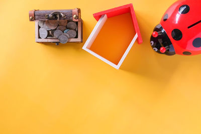 High angle view of coin box with ladybug piggy bank and house model over yellow background