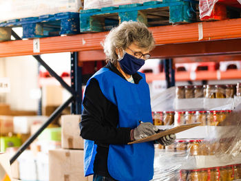 Side view focused female worker in blue vest and face mask taking notes in clipboard while standing near stacks in warehouse