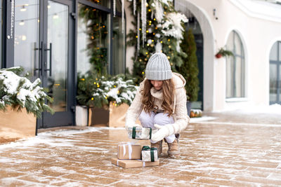 A smiling girl in mittens collects gift boxes from the snow outside in winter
