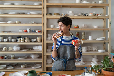 Relaxed woman in apron drink coffee from handmade ceramic cup and has snack in pottery studio