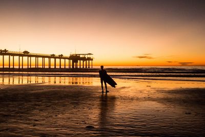 Silhouette man with surfboard standing on shore at beach against sky during sunset