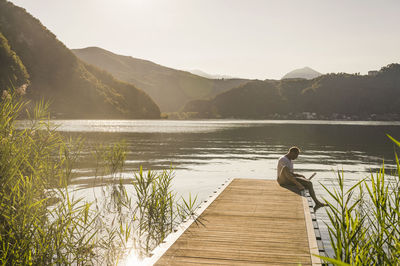 Mature man using laptop on jetty over lake by mountains