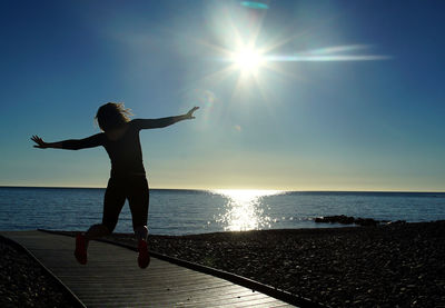 Rear view of woman jumping over boardwalk by sea against sky during sunny day