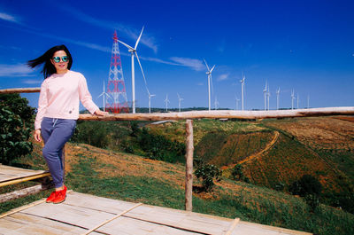 Full length portrait of young woman standing against wind turbines