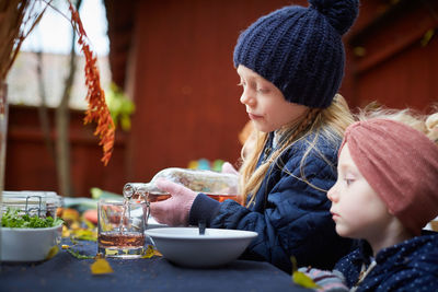 Girl pouring drink in glass while sitting by female sibling at table