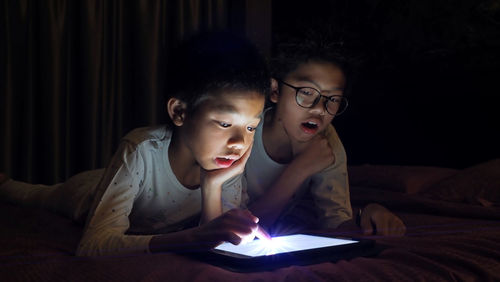 Siblings using digital tablet while lying on bed in darkroom