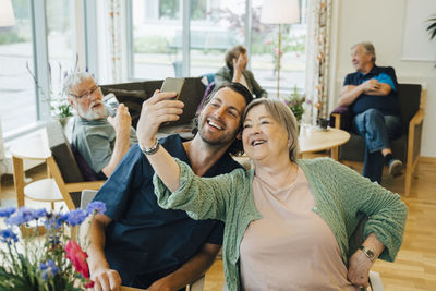 Smiling senior woman taking selfie with male nurse while sitting at retirement home