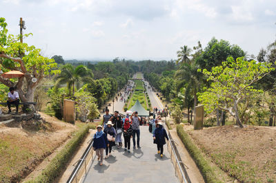 People walking on road amidst plants and trees against sky