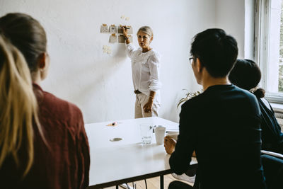 Mature businesswoman writing on adhesive note while explaining colleagues during seminar in office