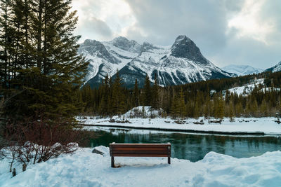 Park bench surrounded by snow and pines overlooking river and snowcapped mountains and dramatic sky.