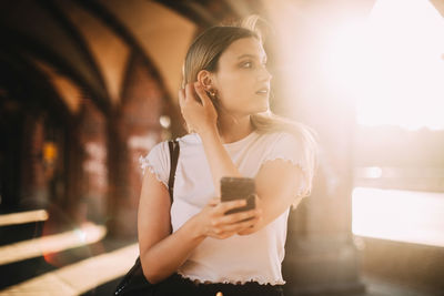 Young woman looking away while holding mobile phone on footpath in city during sunny day