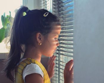 Side view of girl looking through window while standing at home