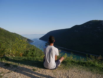 Rear view of man sitting on mountain against clear sky