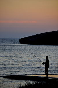 Silhouette man fishing on shore at beach against sky during sunset