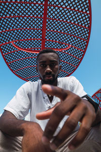 From below of african american male sitting in street on background of blue sky and pointing finger at camera