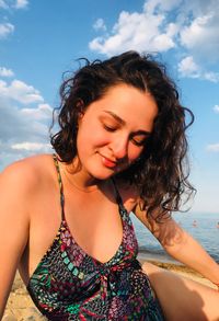 Smiling young woman wearing swimsuit sitting at beach against sky