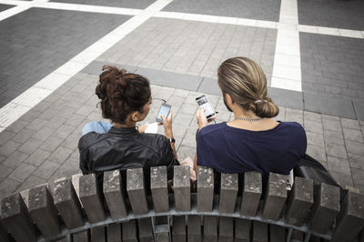 High angle view of man showing mobile phone to friend while sitting on wooden bench in city