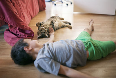 Little boy playing with cat while lying on hardwood floor at home