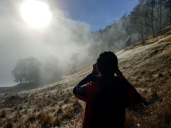 Rear view of young men photographing snow-covered foggy forest against sunlight