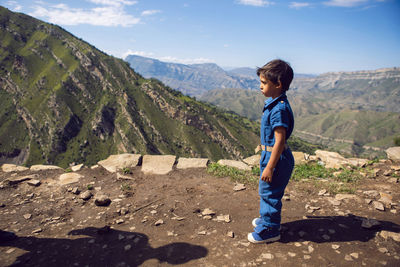 Disgruntled child is a traveler boy in a blue jumpsuit standing in the mountains in summer