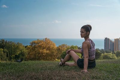 Young woman sitting on grass by sea against sky