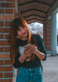 Young woman using phone while standing against brick wall