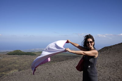 Happy woman holding shawl while standing on mountain against blue sky