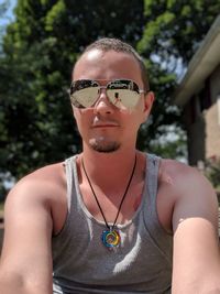 Portrait of young man wearing sunglasses while sitting against trees during sunny day