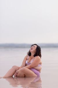 Delighted curvy female in bikini sitting in water of pink pond in summer and looking up