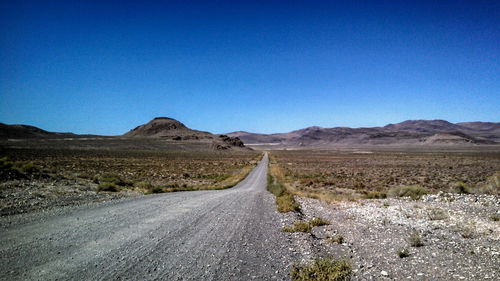 Road amidst field leading towards rocky mountains against clear blue sky