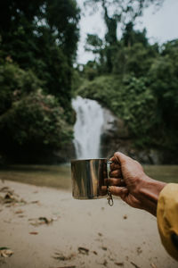 Close up shot of a man's hand holding coffee mug with waterfall in the background.camping.