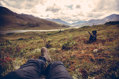 Low section of man lying down with dog on grass and mountains in background