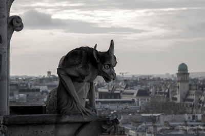 Statue of gargoyle on building in city against cloudy sky during sunset