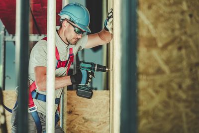 Side view of man drilling wall at construction site