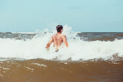 Rear view of shirtless teenage boy enjoying in sea against sky during sunny day