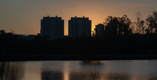 Silhouette buildings by lake against sky at sunset