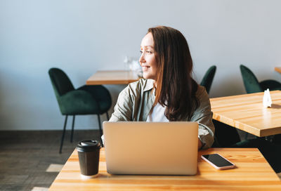 Adult smiling brunette business woman forty years working on laptop using mobile phone in cafe