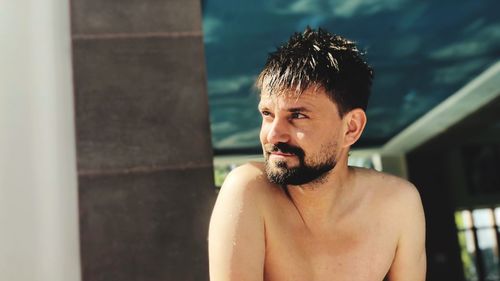 Shirtless man looking away while sitting against house