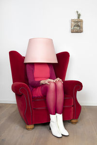 Woman sitting on armchair while face covered with lamp shade